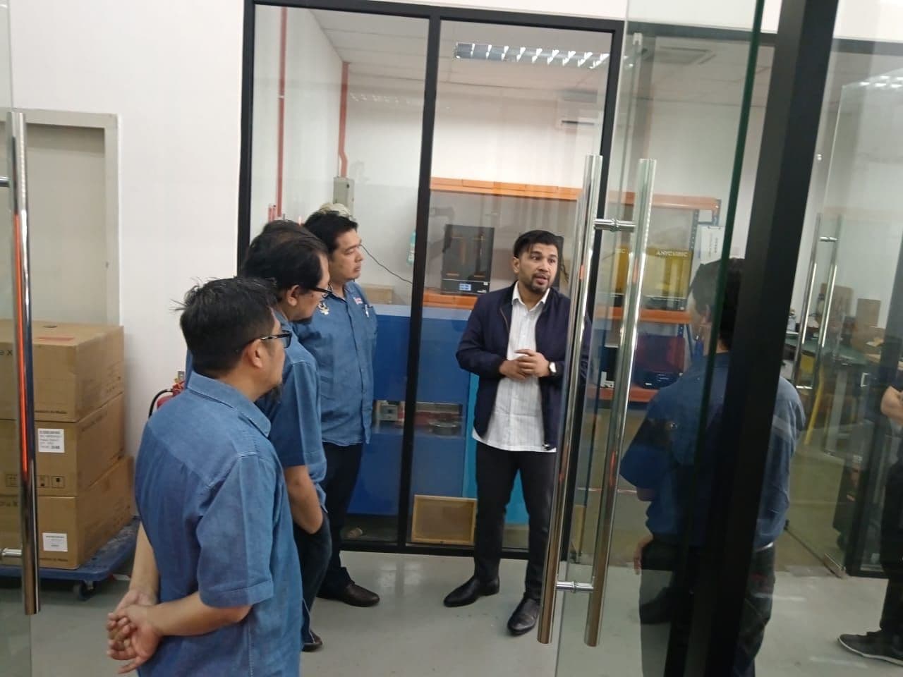 3D Gens representative briefing UPM researchers outside the polymer 3D printing lab, with printers visible through the glass partition